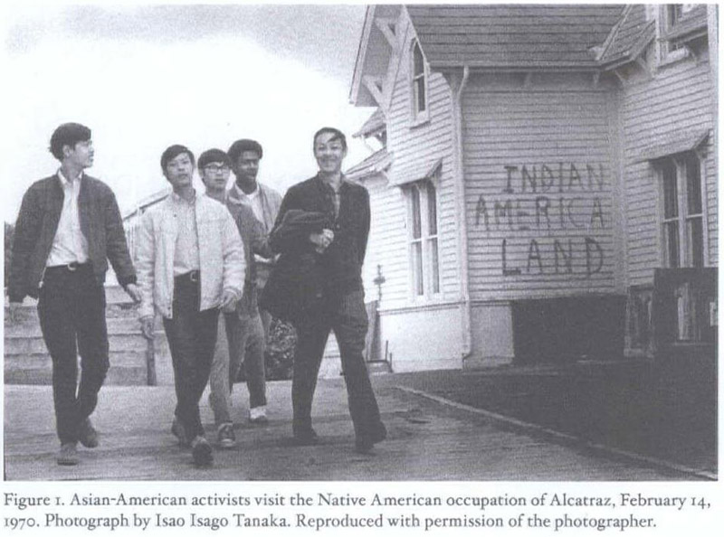 A group of predominantly East Asian men stroll confidently down a street. Graffiti on a house beside them reads 'Indian American Land'. the picture depicts an Asian American delegation to the occupation of Alcatraz in 1970.