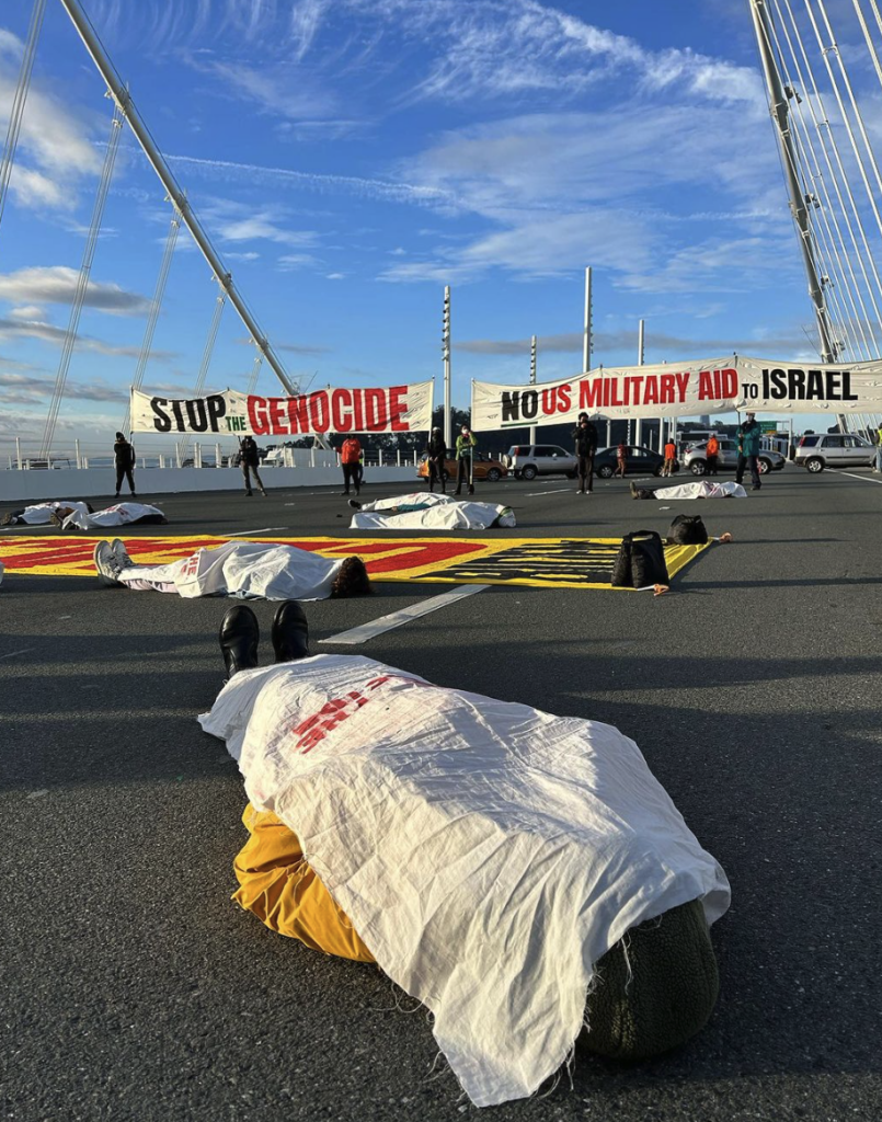 A group of people lies on the street of the Bay Bridge in San Francisco with a white fabric draped over each body. Behind them are large banners that read "Stop the Genocide" and "No US Military Aid to Israel".